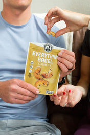 Person holding a bag of Everything Bagel Cashews with another person's hand reaching in to grab a nut.