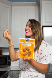 Woman holding a bag of Honey Roasted Chipotle Peanuts in a kitchen tossing peanut into mouth