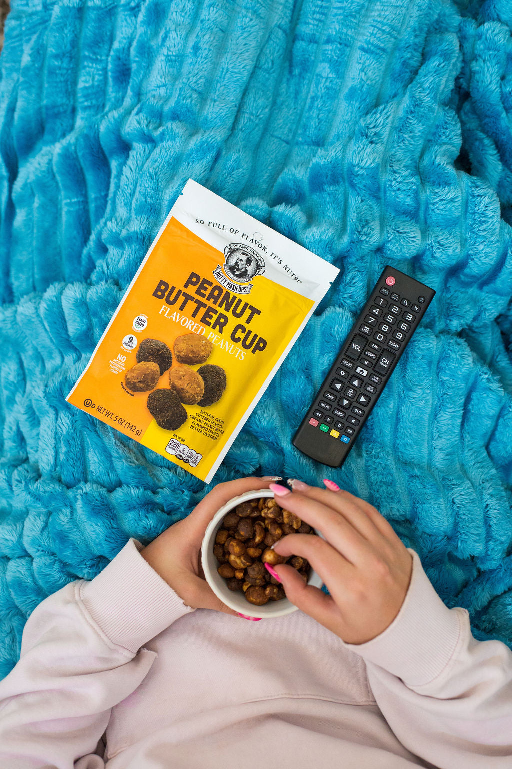 Person holding a bowl of nuts next to a box of peanut butter cups and a remote control on a blue blanket.