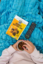 Person holding a bowl of nuts next to a box of peanut butter cups and a remote control on a blue blanket.