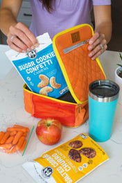 Person holding a bag of sugar cookie confetti cookies putting it into a lunchbox with a blue tumbler and snacks on a table.