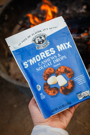 Hand holding a package of s'mores cashex mix bagwith a campfire in the background