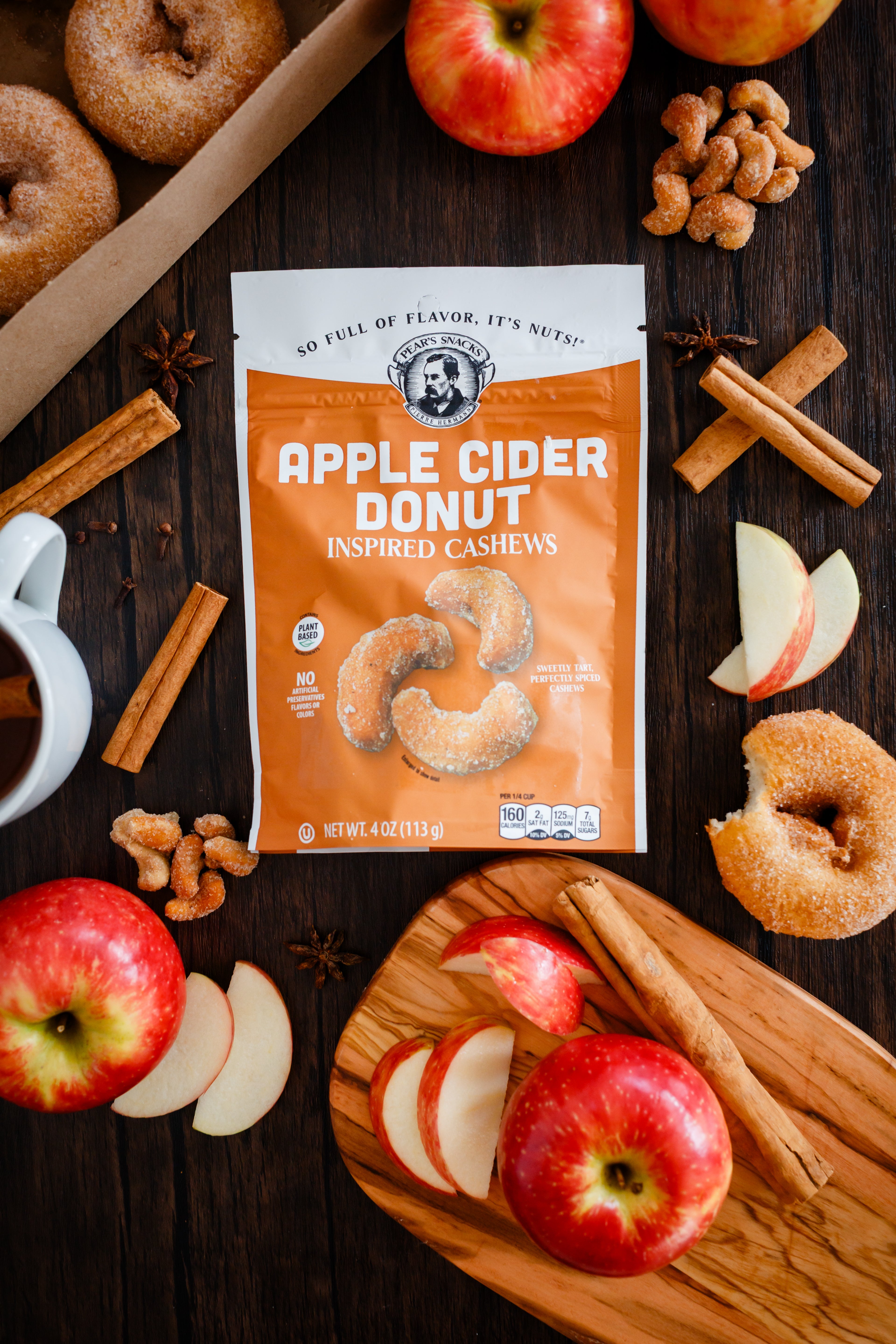 Bag of Apple Cider Donut Inspired Cashews on a wooden table with apples, donuts, and cinnamon sticks.